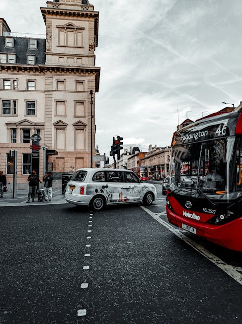 A busy urban street scene showing a white taxi with advertising graphics parked near the curb, partially blocking a pedestrian crossing with dashed white lines. To the right, a red and black double-decker bus displaying route information and the destination 'Paddington' is stopped at a bus stop, with its front window reflecting surrounding buildings. In the background, a corner building with ornate architecture and beige stonework rises above the street, featuring multiple windows and decorative detailing beneath a dark roof. Several pedestrians are visible on the sidewalk waiting to cross or engaged in conversation, some with backpacks or shopping bags. The scene is illuminated by natural daylight under an overcast sky, with some traffic lights visible and a hint of nearby street signage. The overall atmosphere suggests a typical city intersection, where private transport and public transit intersect, emphasizing the importance of efficient rubbish collection and waste management around busy urban areas, which companies such as House Clearance Paddington support through independent waste removal services.