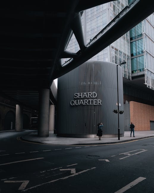 A modern urban scene showing a curved building with vertical gray metal cladding that features the signage 'SHARD QUARTIER' in metallic letters. The building is situated at the corner of a paved street with road markings and black metal lampposts positioned nearby. Tall glass office buildings with a blue tint rise in the background, reflecting the sky and surrounding structures. Overhead, a dark steel canopy with large supporting beams extends across the street, providing shelter and structural support for the surrounding architectural elements. Two pedestrians are visible walking on the sidewalk next to the building, one dressed in dark clothing with a backpack, the other in a light-colored coat. The scene is softly lit, with natural daylight illuminating the urban environment and the sleek, contemporary design of the buildings, creating a professional atmosphere typical of cityscapes with mixed-use developments and potential private waste handling facilities associated with commercial properties like the one shown, highlighting the importance of proper rubbish management in urban areas supported by services like House Clearance Paddington.
