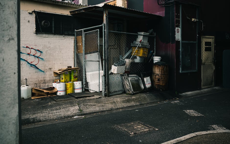 A collection of various waste items stored outdoors in a small, fenced enclosure attached to a building, situated on a paved street. The waste includes multiple white and yellow plastic buckets, some stacked on top of each other, and cardboard boxes placed on the ground. Inside the enclosure, there are several trays, containers, and a yellow plastic crate, along with a mop or broom leaning against the fencing. Nearby, a wooden barrel and a white basin are visible, with additional items stacked and leaning against the wire mesh fencing. The surrounding environment features a brick wall with graffiti to the left, and the building on the right has a dark door and window with barred security screens. The scene is lit by natural daylight, highlighting the textures of the waste materials, metal fencing, and nearby pavement, reflecting elements of private waste storage that might be addressed through professional rubbish removal services such as those offered by House Clearance Paddington.