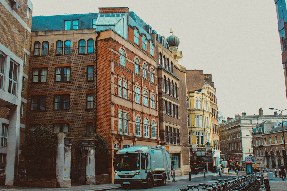 A street scene in an urban area showing a white waste collection truck parked on the right side of the road in front of multi-story buildings with diverse architectural styles. The building closest to the foreground has a reddish-brown brick facade with large, arched windows framed by decorative stonework, and a sloped, teal-colored roof with small dormer windows. Adjacent buildings on either side feature lighter colors, such as cream and pale yellow, with elaborate window and balcony details. The street is lined with bollards and bike racks, and a few pedestrians can be seen further down the sidewalk. The environment is illuminated by natural daylight, creating a clear and neutral atmosphere. The presence of the waste collection vehicle, typical for private rubbish removal or on-site clearance, subtly relates to the context of waste management services provided by House Clearance Paddington in the W2 area, emphasizing the urban scope of private rubbish handling and alternative disposal methods.