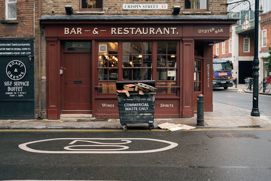 A black commercial waste bin labeled 'COMMERCIAL WASTE ONLY' positioned on a city sidewalk in front of a closed restaurant with a maroon exterior. The bin is partially filled with cardboard boxes and paper, with some discarded items spilling onto the pavement nearby. The restaurant features large glass windows divided into multiple panes, a sign indicating it is a bar and oyster restaurant, and decorative wooden trim painted in a matching maroon color. Adjacent to the bin, a white-painted street marking is visible on the asphalt. To the left, a blackboard sign advertises a self-service buffet, and to the right, a black bollard and a lamppost are present, with vehicles and bicycles seen further down the street. The scene appears during daylight, with natural light illuminating the environment, and the overall setting suggests an urban area where private waste collection or on-site clearance might be needed as part of rubbish removal services offered by House Clearance Paddington.