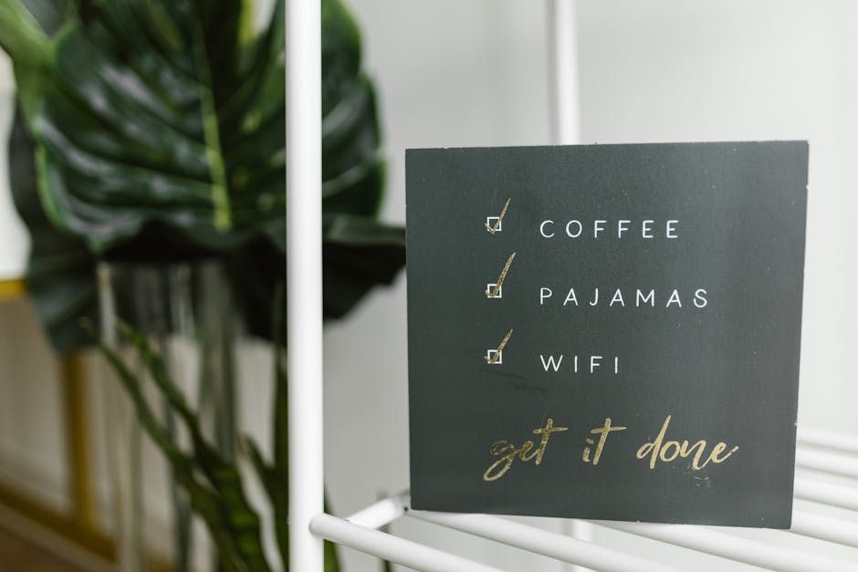 A close-up view of a black chalkboard-style sign with a list of tasks featuring white and gold lettering, placed on a white metal stand. The list includes checkboxes next to the words 'COFFEE,' 'PAJAMAS,' and 'WIFI,' with two of the checkboxes marked with tick symbols. Behind the sign, there is a large, dark green, glossy-leaved potted plant positioned on a light-colored wooden or tiled floor near a plain light gray or white wall. The environment appears to be indoors, possibly in a modern home or café setting, with natural lighting illuminating the scene. The sign's message, 'get it done,' is written in gold cursive at the bottom, blending utility with decorative style, and subtly connecting to themes of task completion and productivity.