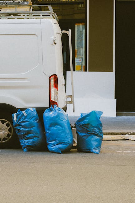 A white commercial van parked on a street curb with its rear door slightly open, showing part of the interior loading area. In front of the van, there are three large blue rubbish bags made of durable plastic, filled with waste, and tightly gathered at the top. The bags are positioned on the pavement, which appears smooth and grey, and they are leaning slightly against the rear wheel of the van. In the background, there is the exterior of a building with a glass door or window reflecting light, and a white architectural element possibly part of a facade or a step. The scene suggests a private or independent waste collection activity, aligned with services like rubbish removal or on-site clearance, often managed by entities such as House Clearance Paddington, to facilitate waste disposal outside of local authority collection. Lighting is neutral, with daylight illuminating the scene clearly and evenly, emphasizing the textures of the plastic bags and the van's surface.