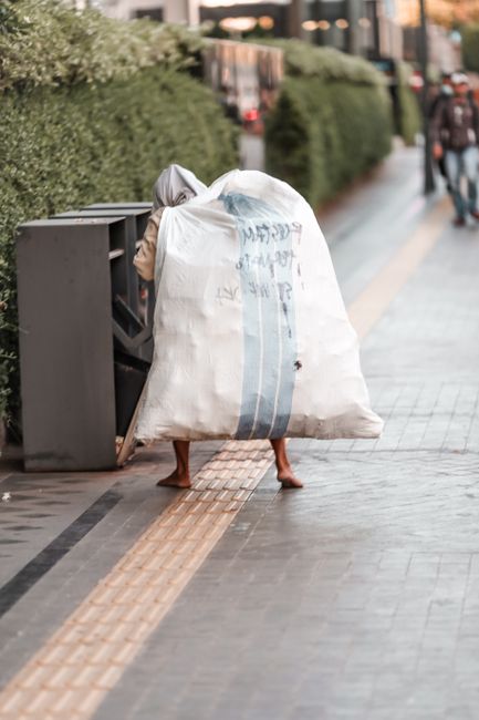A person with their legs visible standing on a paved sidewalk, partially concealed by a large, white plastic sack filled with waste, which covers their upper body. The sack has vertical blue stripes and some illegible text printed on it. The individual appears to be loading or unloading rubbish into or from a black, multi-compartment waste bin positioned beside them. The scene is set outdoors in an urban environment, with a neatly trimmed hedge and residential buildings in the background. The adjacent pavement features tactile paving strips, and a few pedestrians are visible walking further down the street. The scene suggests a typical outdoor rubbish collection or disposal process, with the individual possibly engaged in private waste handling or on-site clearance, an activity often supported by local rubbish removal services such as House Clearance Paddington. The lighting indicates natural daylight, contributing to a clear and neutral visual atmosphere.