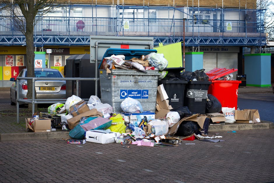 A person with their legs visible standing on a paved sidewalk, partially concealed by a large, white plastic sack filled with waste, which covers their upper body. The sack has vertical blue stripes and some illegible text printed on it. The individual appears to be loading or unloading rubbish into or from a black, multi-compartment waste bin positioned beside them. The scene is set outdoors in an urban environment, with a neatly trimmed hedge and residential buildings in the background. The adjacent pavement features tactile paving strips, and a few pedestrians are visible walking further down the street. The scene suggests a typical outdoor rubbish collection or disposal process, with the individual possibly engaged in private waste handling or on-site clearance, an activity often supported by local rubbish removal services such as House Clearance Paddington. The lighting indicates natural daylight, contributing to a clear and neutral visual atmosphere.