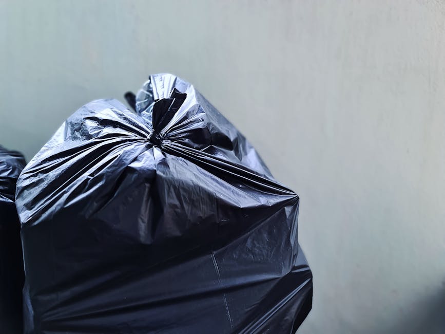 Close-up of several black plastic rubbish bags tightly sealed with twist ties, placed on a surface near a plain light-colored wall. The bags are made of glossy plastic, reflecting light on their smooth, slightly crinkled surfaces. In the background, additional bags are partially visible, stacked together, indicating a collection of waste awaiting removal. The environment appears to be an outdoor area or driveway, suitable for private rubbish collection services such as those offered by House Clearance Paddington. The scene suggests an organized effort to manage waste through alternative disposal methods, avoiding local authority rubbish collection, and highlights the presence of privately managed rubbish removal in a residential setting. Lighting is neutral, with no strong shadows, emphasizing the utilitarian purpose of the scene.