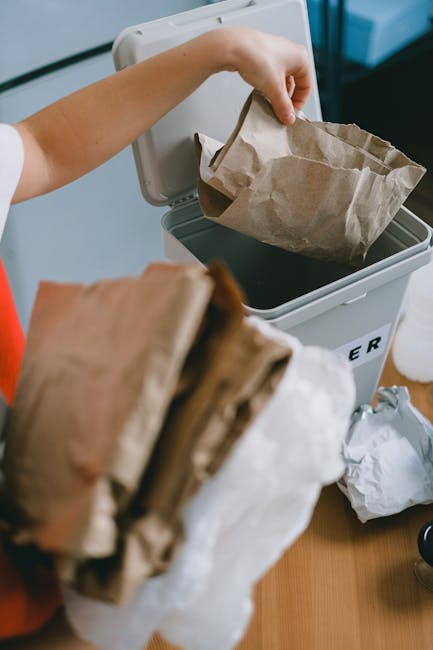 A person’s hand is shown lifting a crumpled brown paper bag out of an open grey rubbish bin, which has a white lid and is positioned on a wooden surface. Inside the bin, a similar paper bag is visible, filled with dark waste material, possibly food or general rubbish. In the foreground, a bundle of additional crumpled paper bags is held in another hand, partially obscuring the view. The background includes a light-colored wall and part of a countertop or desk, indicating an indoor environment. This scene relates to private waste disposal and alternative waste handling methods such as on-site rubbish collection, which is a service some companies, like House Clearance Paddington, provide for area residents. The overall setting emphasizes a neat, organized approach to rubbish collection, reflecting professional waste management practices suitable for house clearance or individual rubbish removal in a domestic context.