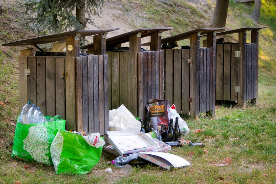 Close-up of several black plastic rubbish bags tightly sealed with twist ties, placed on a surface near a plain light-colored wall. The bags are made of glossy plastic, reflecting light on their smooth, slightly crinkled surfaces. In the background, additional bags are partially visible, stacked together, indicating a collection of waste awaiting removal. The environment appears to be an outdoor area or driveway, suitable for private rubbish collection services such as those offered by House Clearance Paddington. The scene suggests an organized effort to manage waste through alternative disposal methods, avoiding local authority rubbish collection, and highlights the presence of privately managed rubbish removal in a residential setting. Lighting is neutral, with no strong shadows, emphasizing the utilitarian purpose of the scene.