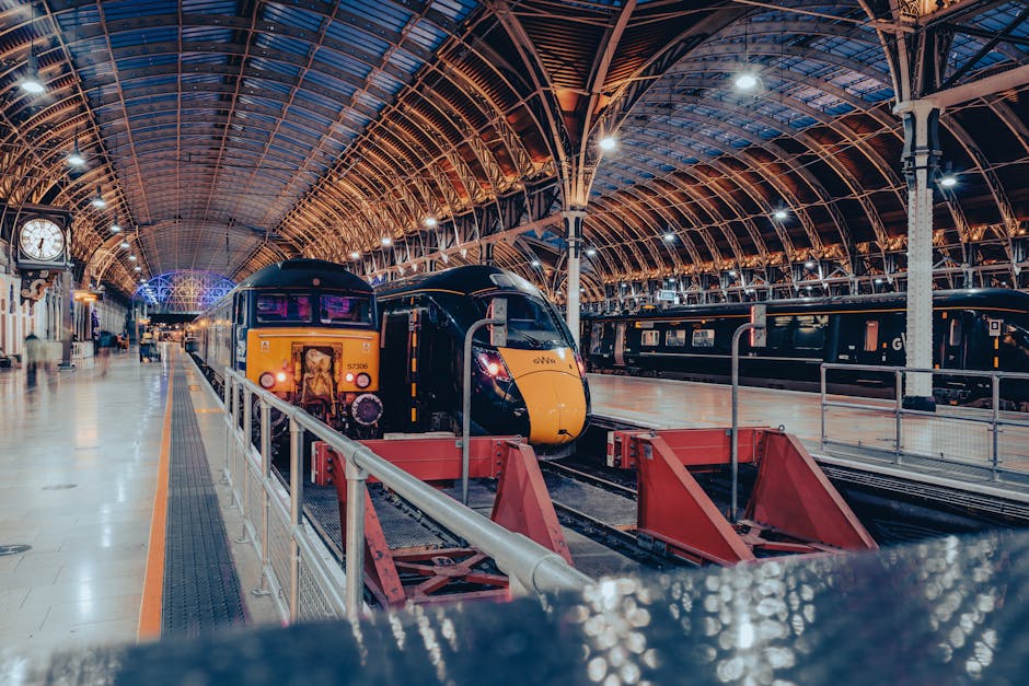 A modern, streamlined yellow and black train resting on railway tracks inside an expansive, historic train station with an ornate iron and glass arched ceiling illuminated by bright overhead lights. The platform area features a polished, beige tiled floor and a white and black clock mounted on a decorative column to the left. To the left of the train, there is a yellow-striped safety barrier; behind it, some passengers and station staff are visible, adding subtle activity to the scene. The station environment includes additional train cars parked along the tracks in the background, and the overall lighting accentuates the warm tones of the metal framework contrasted with the cool blue and purple hues of the station’s architectural elements. This setting exemplifies the infrastructure used for rail travel and simple railway operations, similar in context to locations where independent waste collection services, such as those provided by House Clearance Paddington, might operate nearby for alternative disposal solutions related to transit or station areas. Visible surface textures include the smooth finish of the train’s exterior, the intricate ironwork of the station structure, and the reflective quality of the tiled floor, all contributing to a clear, professional depiction of a busy railway hub.