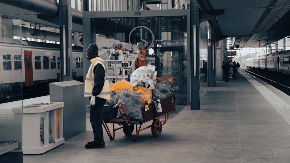 A man dressed in dark clothing, including a beanie and gloves, stands next to a large red wagon loaded with multiple black, grey, and orange rubbish bags, along with some loose debris and smaller containers, situated on a paved platform at a modern train station. The surrounding environment features a glass shelter behind the man, with machinery and equipment visible behind the glass, indicating a possible area for waste collection or storage. To the left, there is a grey waste bin or recycling container, and in the background, train tracks extend into the distance, with another train visible on the right side of the platform. The station's sleek, metallic, and glass architectural elements are prominent, with a few other passengers seen further along the platform. The lighting is natural and diffused, typical of an overcast day, contributing to a neutral, professional atmosphere that emphasizes the scene's focus on waste handling within an urban transit setting, reflecting practices related to independent collection or street-level rubbish removal which may be associated with private waste disposal services like those offered by House Clearance Paddington.