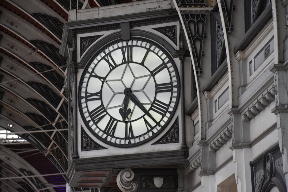 A large, ornate clock mounted on the exterior wall of a historic building, featuring a white face with bold black Roman numerals and black hands, with the time showing approximately 5:25. The clock’s frame is made of dark metal with intricate decorative elements. The building’s facade surrounding the clock is constructed of stone or plaster with detailed moldings and carvings, painted in a light, neutral tone. To the left, part of a modern steel or iron structure with a curved, lattice-like design is visible, suggesting the scene is at a railway or train station. The environment is well-lit with natural daylight, highlighting the texture of the materials and contrasting the classic architectural features with the contemporary structural elements nearby. This scene exemplifies the blend of historical architecture and modern infrastructure, relevant to urban settings that may require frequent building maintenance or renovation, often involving rubbish removal services like those provided by House Clearance Paddington.