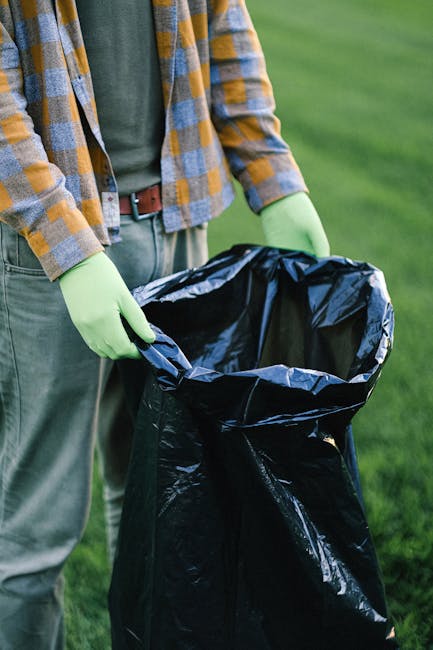 A man dressed in dark clothing, including a beanie and gloves, stands next to a large red wagon loaded with multiple black, grey, and orange rubbish bags, along with some loose debris and smaller containers, situated on a paved platform at a modern train station. The surrounding environment features a glass shelter behind the man, with machinery and equipment visible behind the glass, indicating a possible area for waste collection or storage. To the left, there is a grey waste bin or recycling container, and in the background, train tracks extend into the distance, with another train visible on the right side of the platform. The station's sleek, metallic, and glass architectural elements are prominent, with a few other passengers seen further along the platform. The lighting is natural and diffused, typical of an overcast day, contributing to a neutral, professional atmosphere that emphasizes the scene's focus on waste handling within an urban transit setting, reflecting practices related to independent collection or street-level rubbish removal which may be associated with private waste disposal services like those offered by House Clearance Paddington.