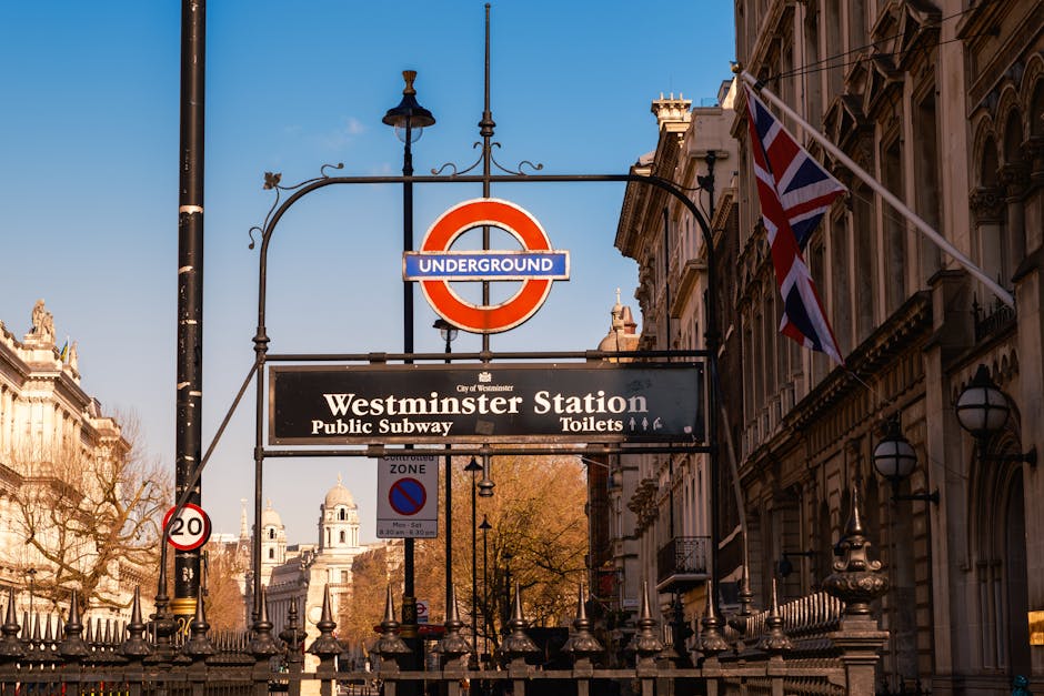 A street view outside Westminster Underground Station in London, featuring the iconic red and blue London Underground roundel sign suspended on an ornate black wrought iron bracket. Below the sign, there is a black rectangular board displaying white text indicating 'Westminster Station' with directions to the public subway and toilets. To the right, a Union Jack flag is attached to the historic stone façade of a nearby building, showcasing classical architectural details such as decorative cornices and arched windows. The background includes leafless trees and classic London-style street lamps, with clear blue sky overhead indicating daytime. This scene captures a typical London transport hub environment, ideal for illustrating urban infrastructure and access points for public transportation, yet also relevant to discussions about private waste collection and alternative rubbish removal services conducted near busy historic sites like Westminster. The image's lighting highlights the textures of the building stonework, metal signage, and the patterned pavement surrounding the station entrance, contributing to a professional, neutral scene suitable for accessibility-focused content on rubbish removal and house clearance services in the Westminster area.