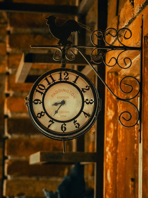 A vintage-style round clock mounted on a decorative wrought iron bracket attached to an exposed brick wall. The clock face features classic black numerals on a cream background, with black hour and minute hands, displaying a time around 7:42. The bracket includes ornamental scrollwork and a silhouette of a bird perched atop the extension arm. The background reveals a dark indoor environment with wooden beams and shelving, contributing to an industrial or rustic aesthetic. The scene captures a sense of timelessness, with the clock's inscription indicating it is from Paddington Station in London, dating back to 1854. This image may relate to how independent or private collection of items, such as vintage or antique pieces, can be part of on-site clearance processes managed by companies like House Clearance Paddington. The detailed focus on the clock and its surroundings aligns with themes of domestic or commercial waste management, emphasizing the importance of careful removal of old or unwanted items in residential or flat clearance settings.
